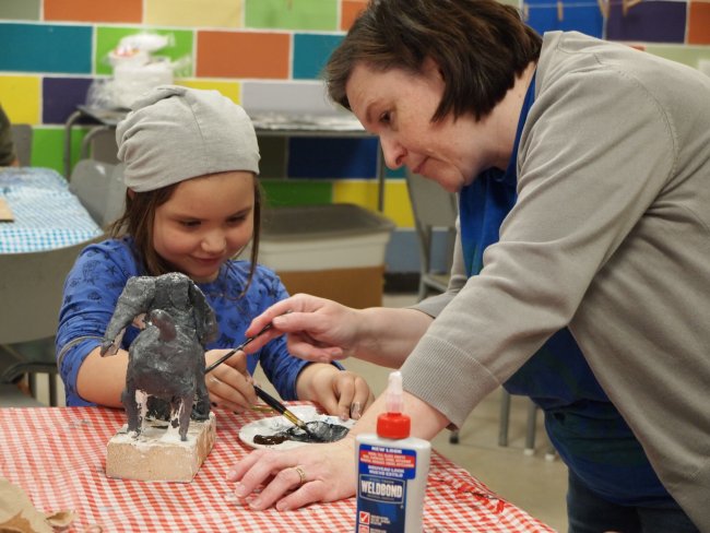 Image of a young child sitting at a studio table painting a sculpture with the help of an adult
