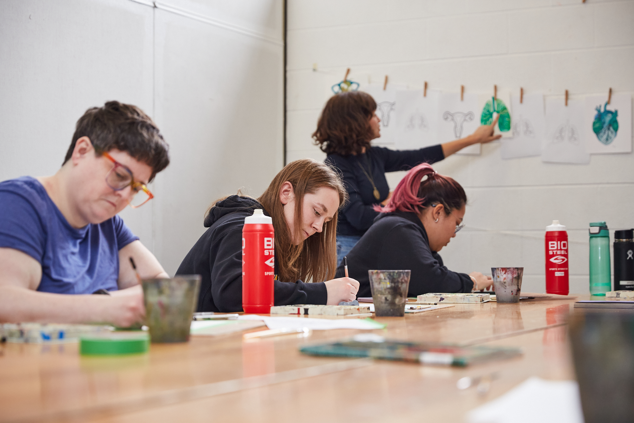 Photo of a group of people sitting around a wooden table. An instructor hangs artwork whilst students sketch on paper.