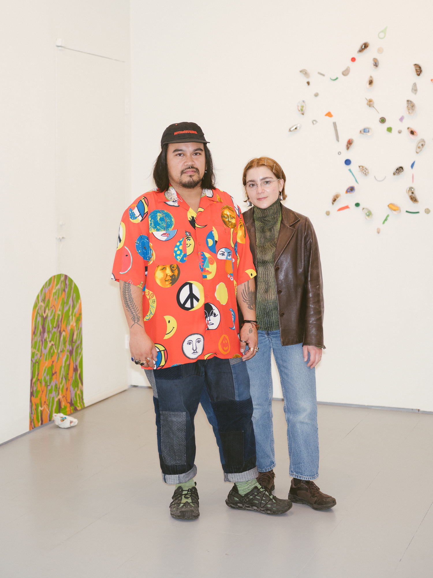 Two people, one male presenting in an orange shirt and jeans, the other, a female presenting person standing on a grey floor