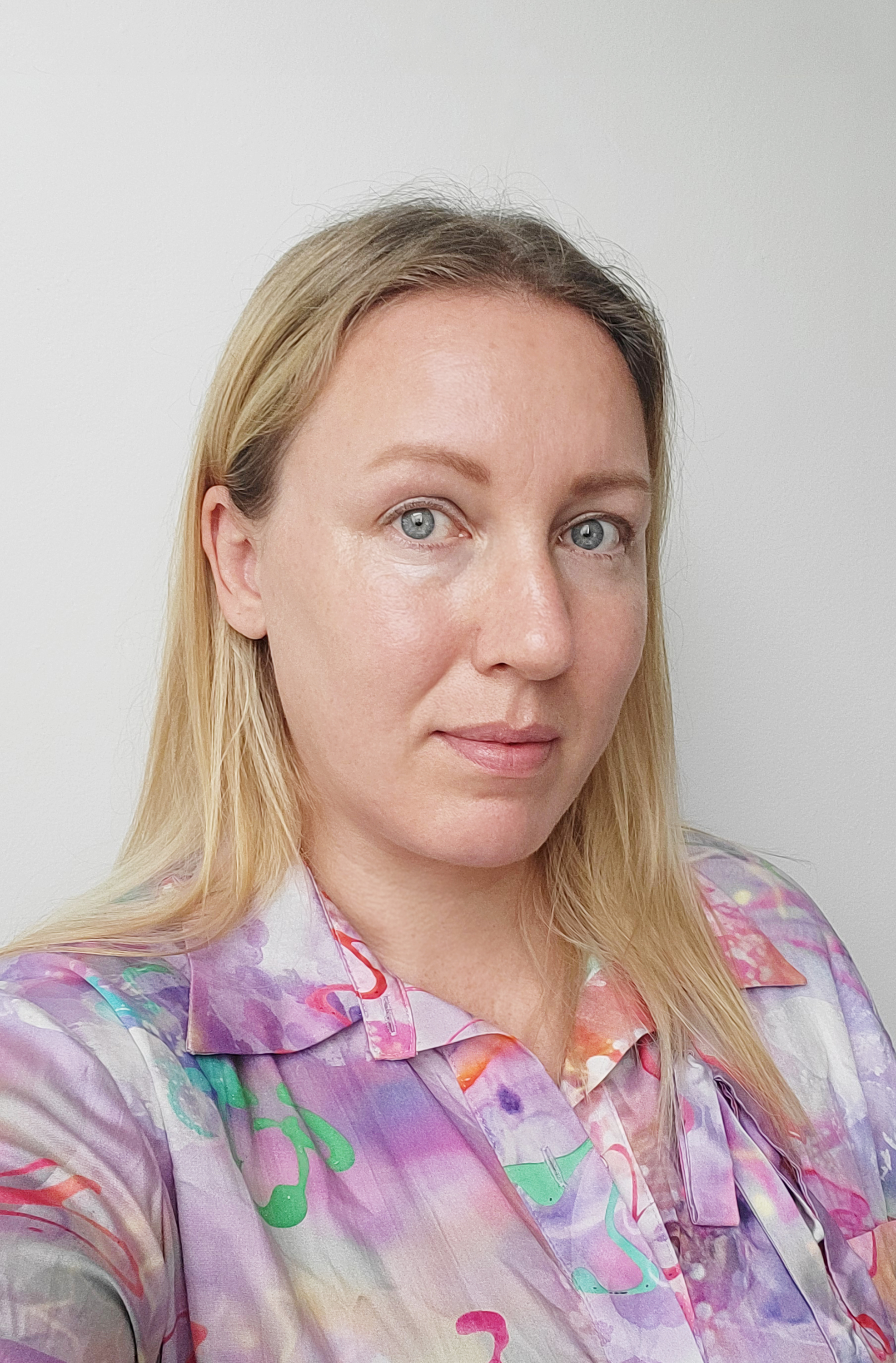 Photo of a Jenine Marsh, a female presenting person with long blonde hair and light blue eyes. She is wearing a multicolored pastel purple and pink shirt with abstract lines of varying lengths and widths