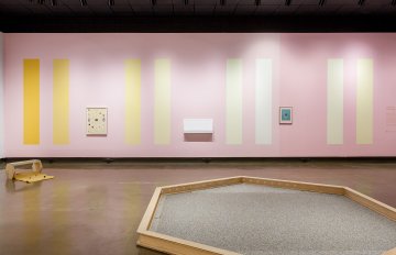 installation shot of gallery exhibition showcasing colourful pastel walls with frames hung on it and a hexagon shape made of wood in the centre