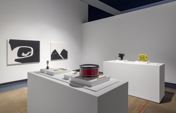 Photo of two plinths holding objects such as a snare drum, taxi sign, taxi person hat. In the background is a black and white painting by Yan Wen Chang of a side view mirror of a car