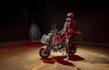 Side view of The Medicine Man sculptural installation of an e-bike rider with an e-bike adorned with many bags, blankets and a life-size mannequin wearing a helmet, mask and winter clothing