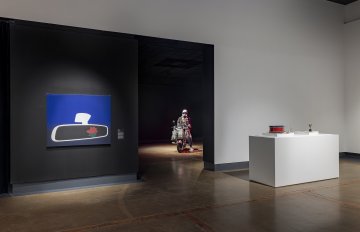 Photo of a black wall with a Yan Wen Chang rearview mirror painting with a rose in the reflection, the back room featuring The Medicine Seller e-bike and mannequin sculpture and museum objects on a white plinth