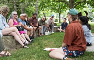 Photo of a group of individuals in a discussion. Some sit on the grassy floor, others sit on rocks.