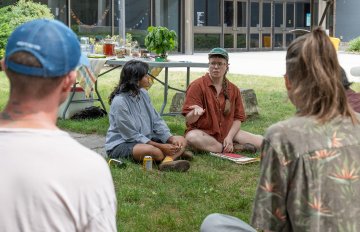 Photo from above of two people sitting on grass in front of a table in front of a few people