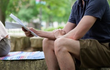 Photo of a young man in shorts holding reading material sitting on a rock in a garden setting