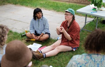 Photo from above of two people sitting on grass in front of a table in front of a few people