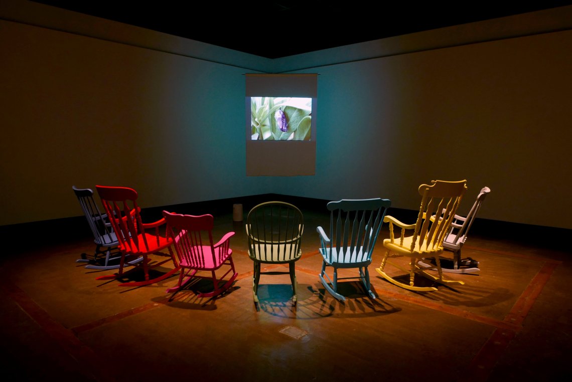 A semi-circle of colourful rocking chairs face a small video projection of a cocoon in a dark gallery