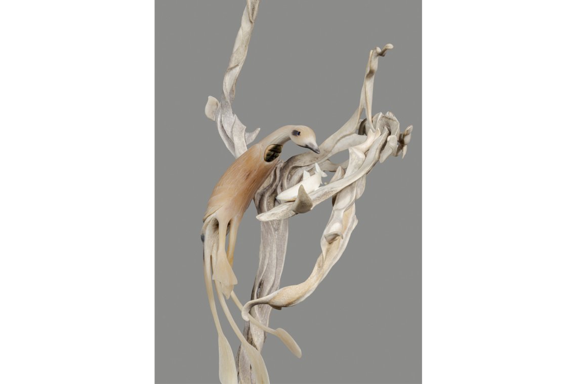 Photo of an antler and ivory sculpture standing on a base of labrodorite. The figure of a loon is leaping in the air to catch a fish.