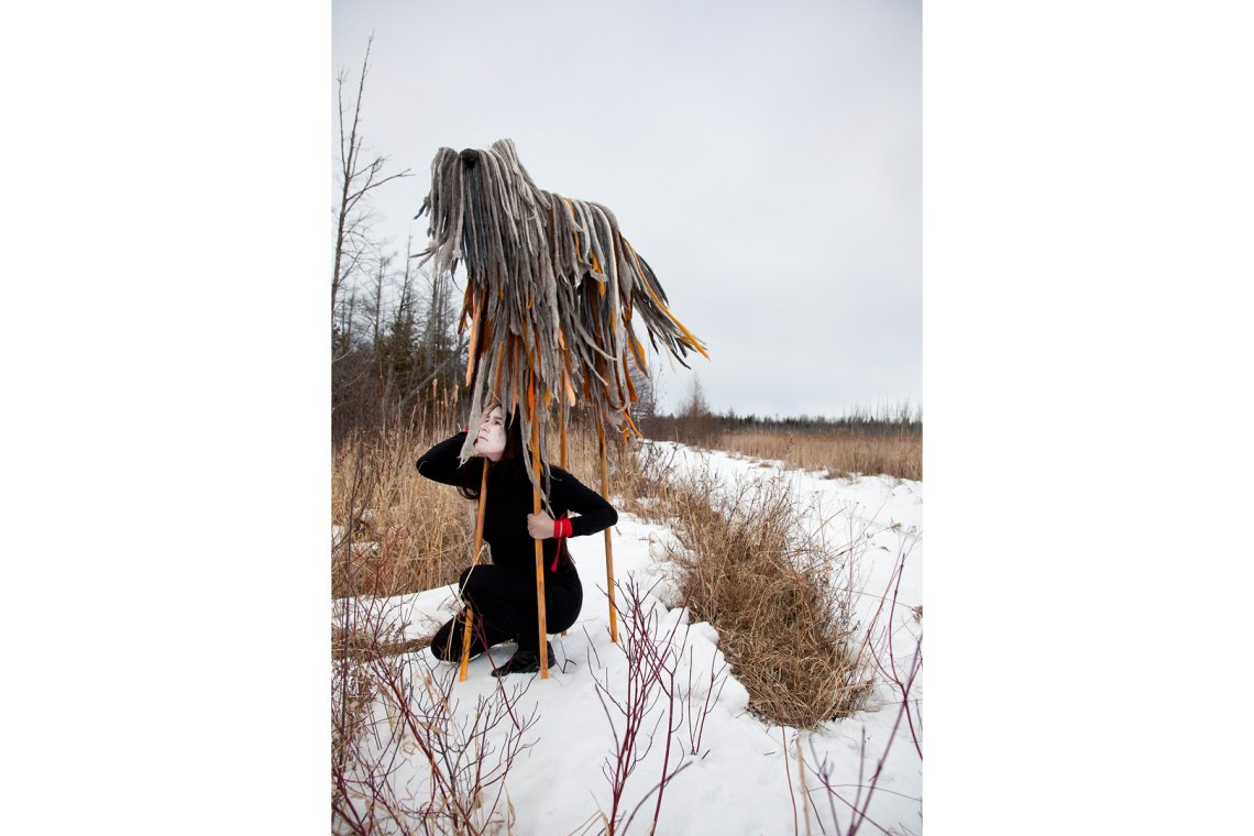 Photo of a person dressed in black with white face paint squatting under four poles holding up various muted colored textured strips of materials