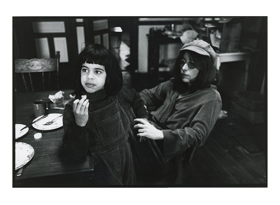 Black and white image of a small child facing away from a camera. An adult sits beside them holding their arm with a blank expression